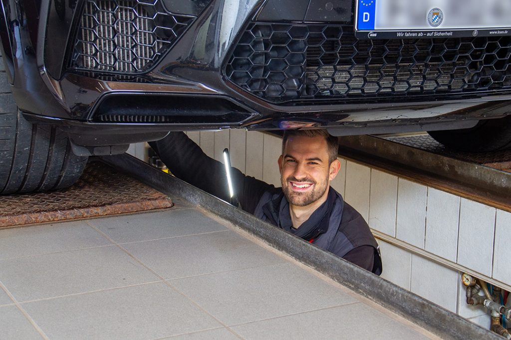 Ein freundlicher Prüfingenieur lächelt unter einem Kfz hervor, während er in der Grube unter dem Fahrzeug eine Prüfung vornimmt.