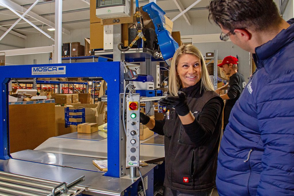 Auf dem Bild ist die Verpackungsabteilung eines Unternehmens mit entsprechender Maschine zu sehen und die Prüfingenieurin des Ing.-Büro Prüfcenter Ebert beim Maschinencheck.